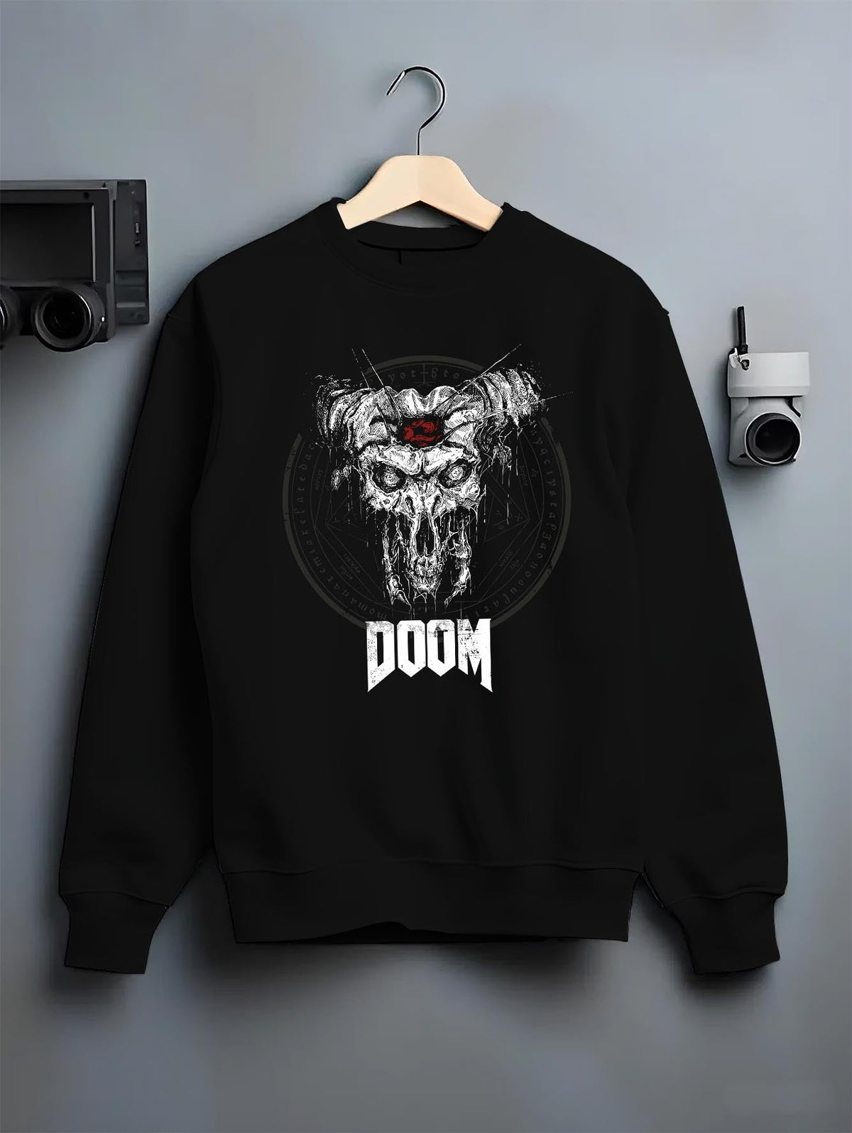 Black crewneck sweatshirt on a hanger against a grey wall, featuring DOOM skull with horns artwork in distressed horror poster aesthetic and retro gaming theme.