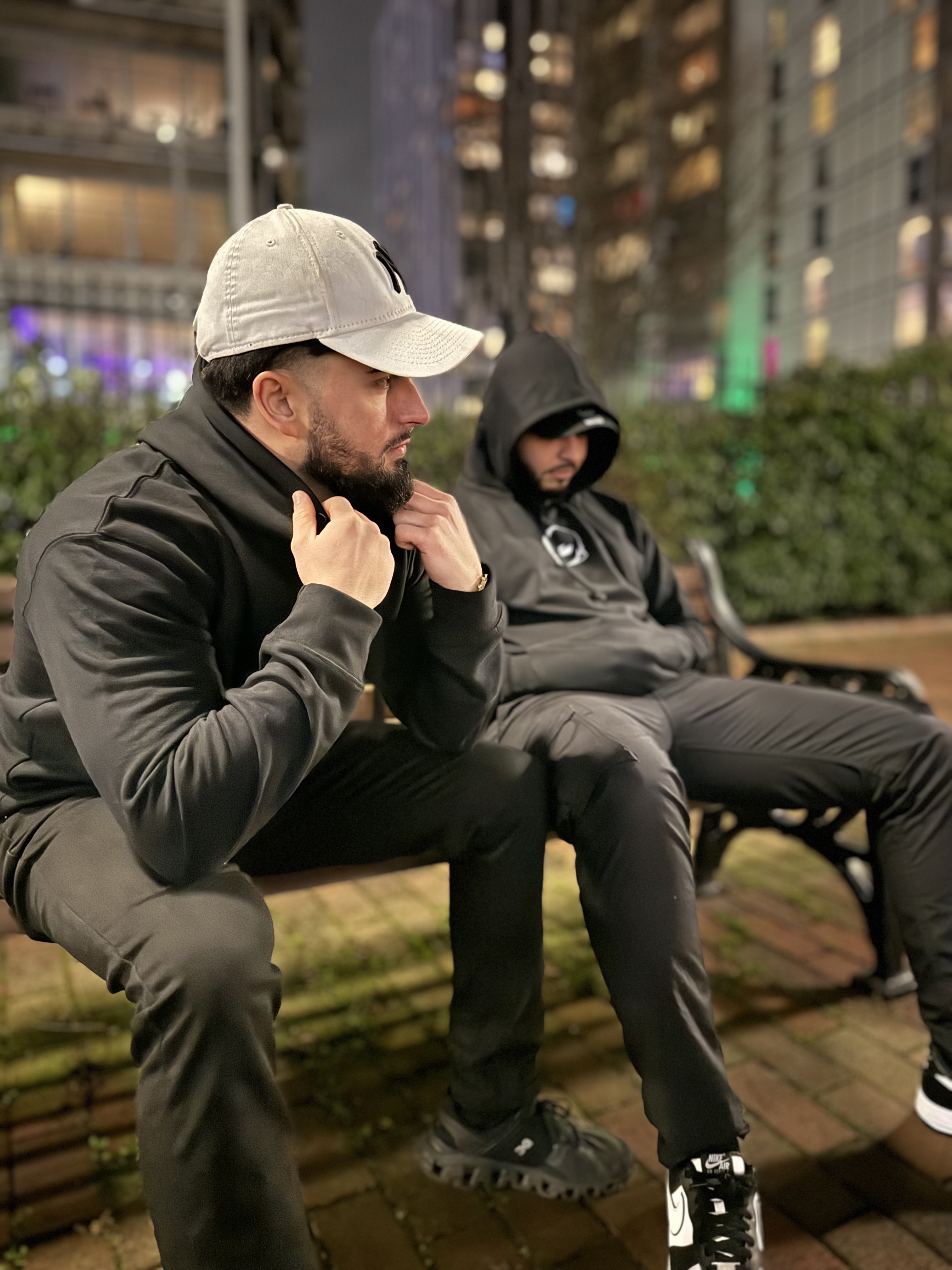 Two men wearing black anime hoodies from Kunai, one featuring Dragon Ball design, streetwear night shoot in cityscape setting for anime fashion fans.