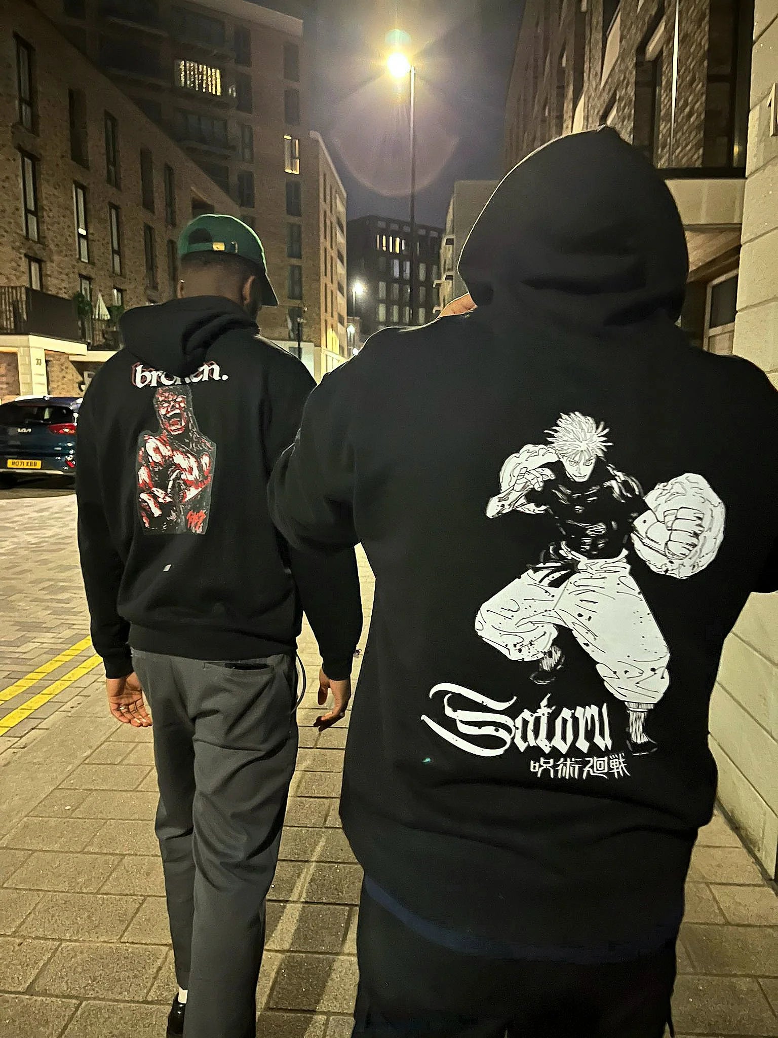 Two people wearing black hoodies with graphic designs on a city street at night.