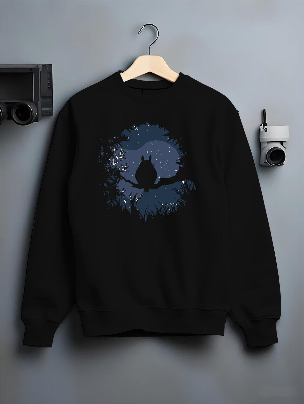 Black crewneck sweatshirt on a hanger against a grey wall, featuring Totoro moonlit forest silhouette, minimalist streetwear back design.