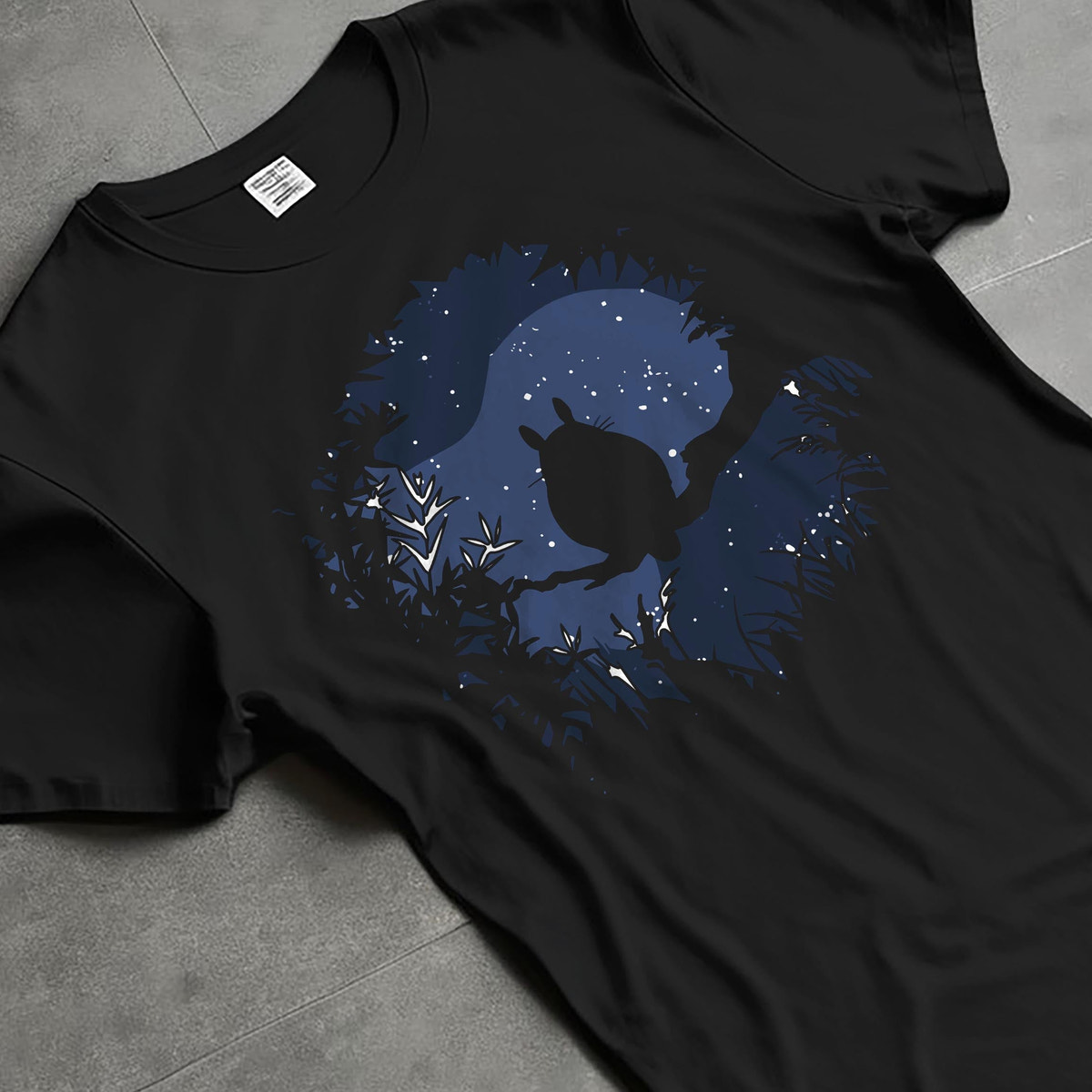 Black T-shirt laid flat with natural fabric folds, featuring My Neighbor Totoro silhouette framed by branches and stars, casual retro graphic.