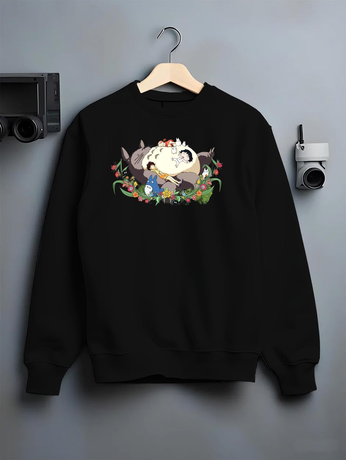 Black crewneck sweatshirt on a hanger against a grey wall, featuring Totoro sleeping surrounded by flowers, nostalgic 90s Japanese animation streetwear.