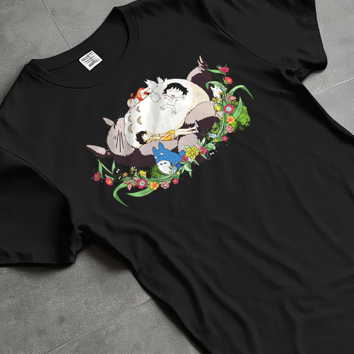 Black T-shirt laid flat with natural fabric folds, featuring Totoro nap tableau and flowers, retro streetwear graphic with vintage anime charm.
