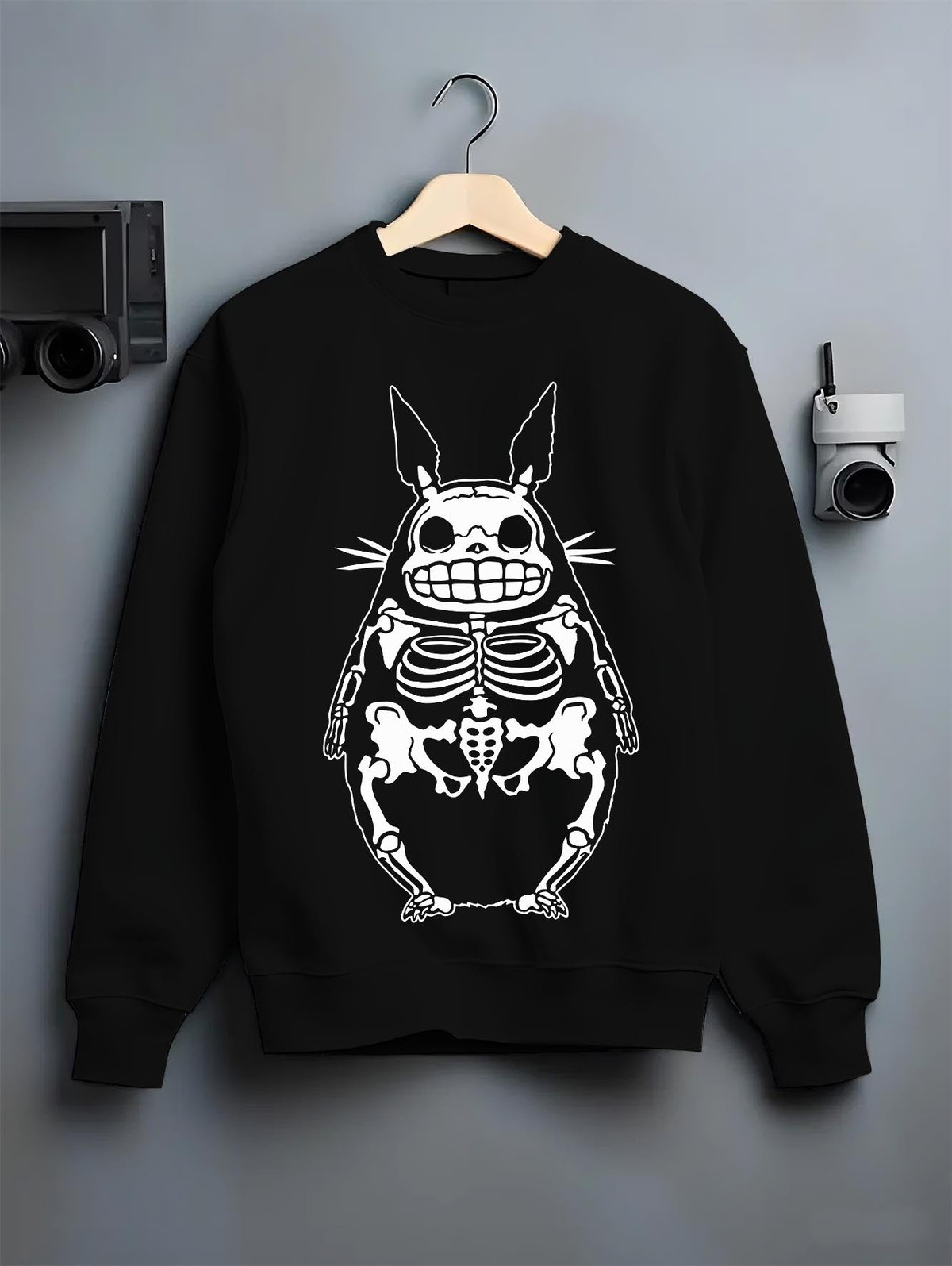 Black crewneck sweatshirt on a hanger against a grey wall, featuring Totoro skeleton x-ray silhouette, retro streetwear graphic with monochrome line art.